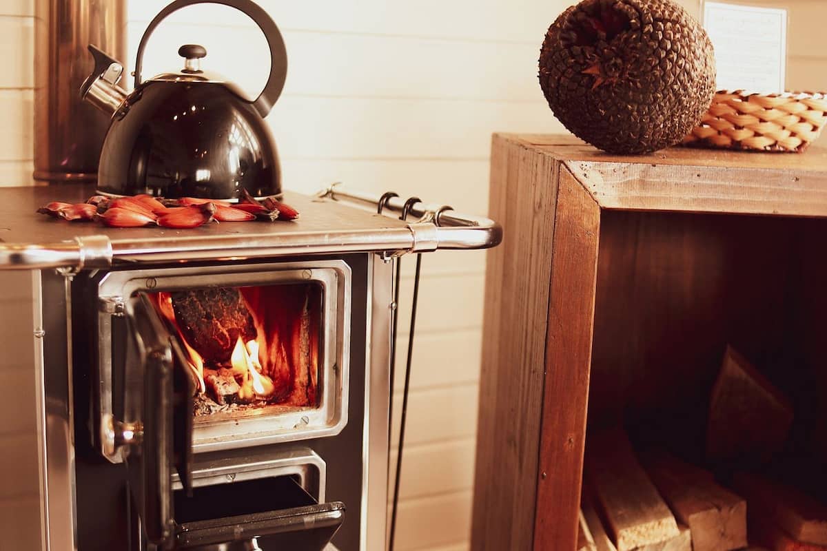 A wood-burning stove with a kettle on top, a fire burning inside, and a stack of firewood beside it.