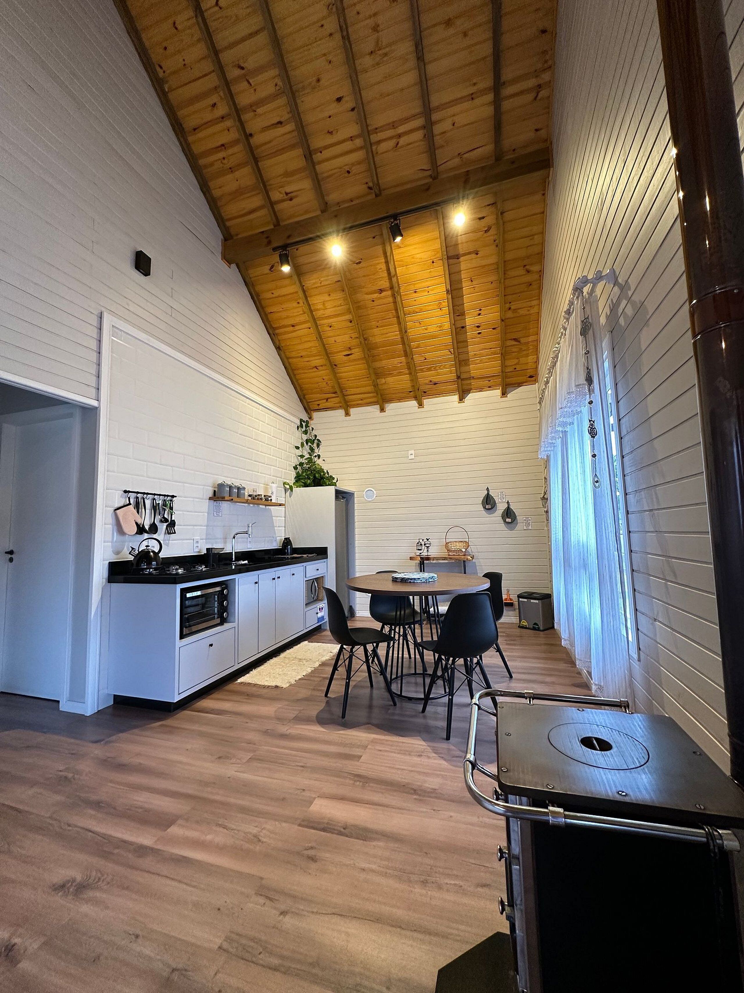 Cozinha moderna com armários brancos, bancada preta e teto de madeira. Há uma área de jantar com mesa e cadeiras, e um forno pequeno embutido nos armários.