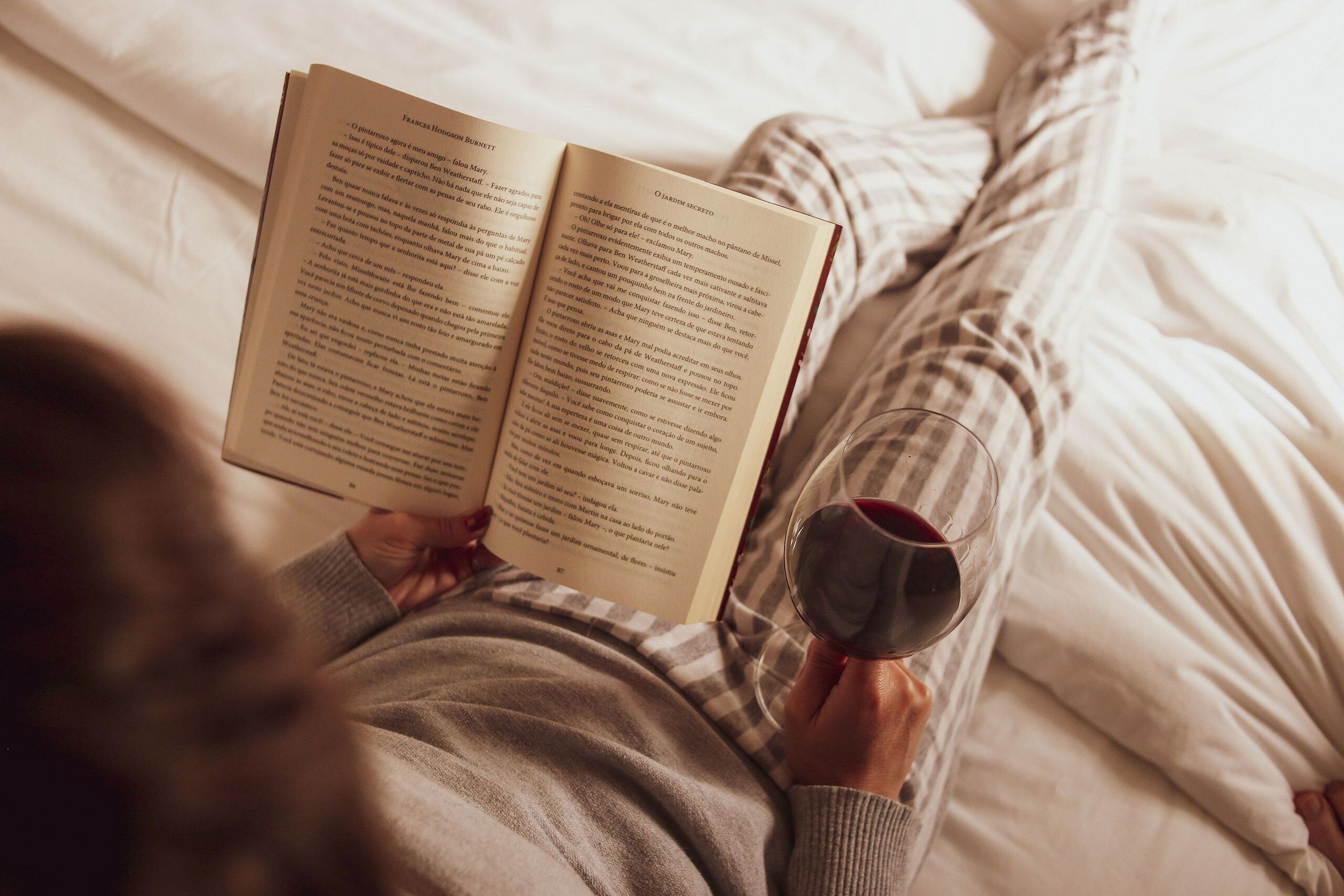 A person lying on a bed reading a book and holding a glass of red wine.