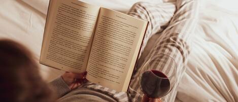 A person lying on a bed reading a book and holding a glass of red wine.
