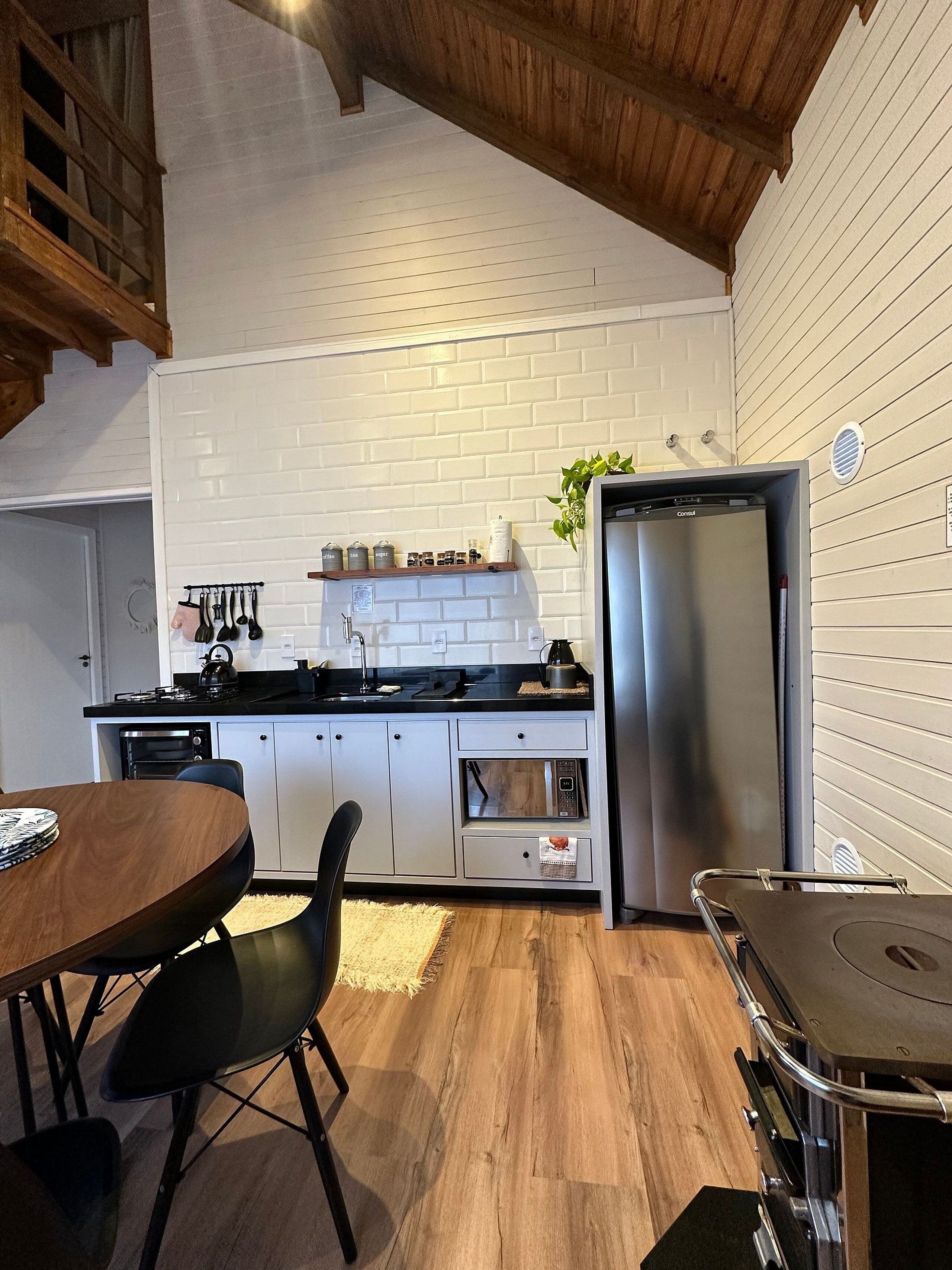 A compact kitchen with white brick walls, a stainless steel refrigerator, and a dining area with a wooden table and black chairs.
