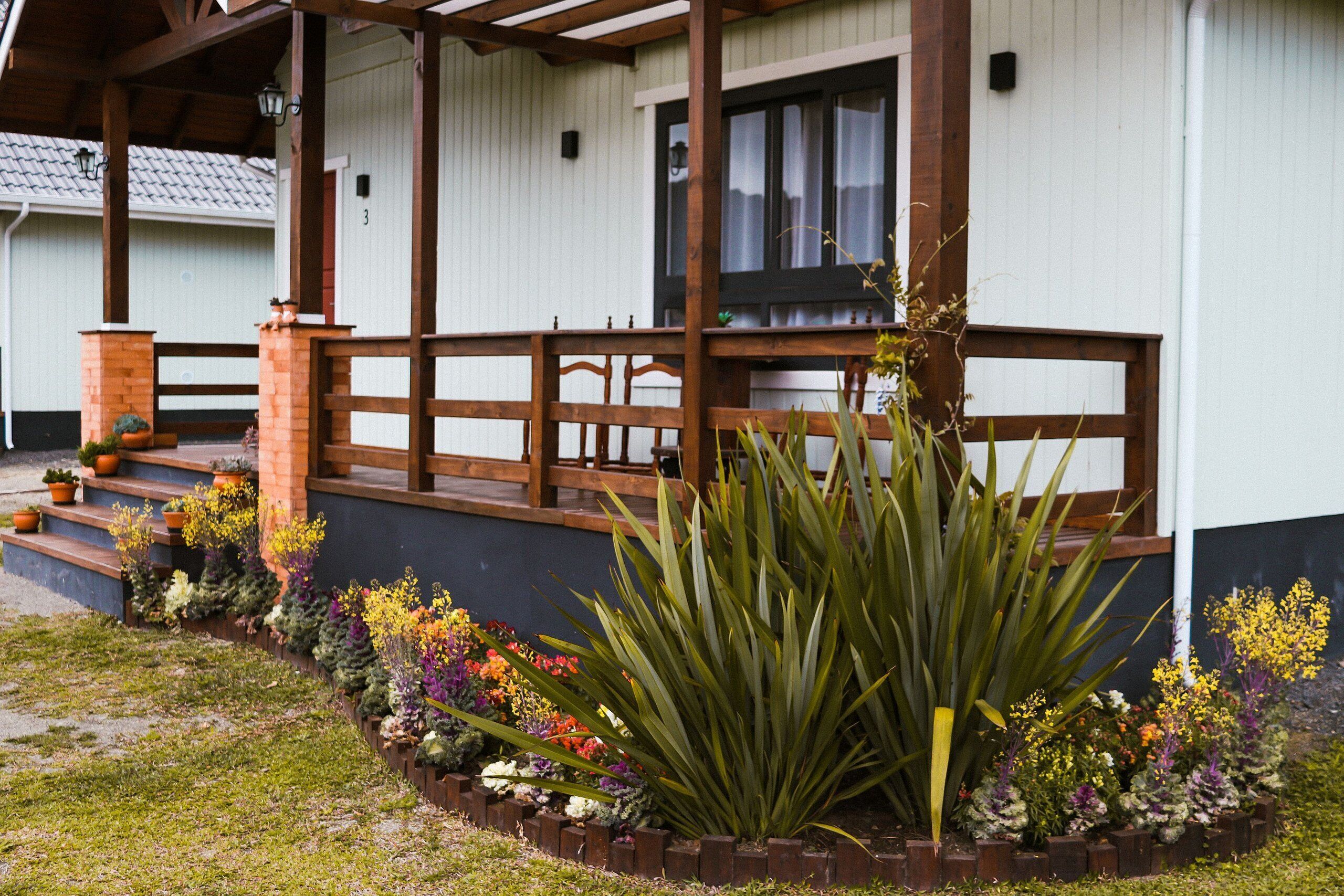 Um edifício com varanda de madeira, degraus até uma porta e um jardim com diversas plantas.