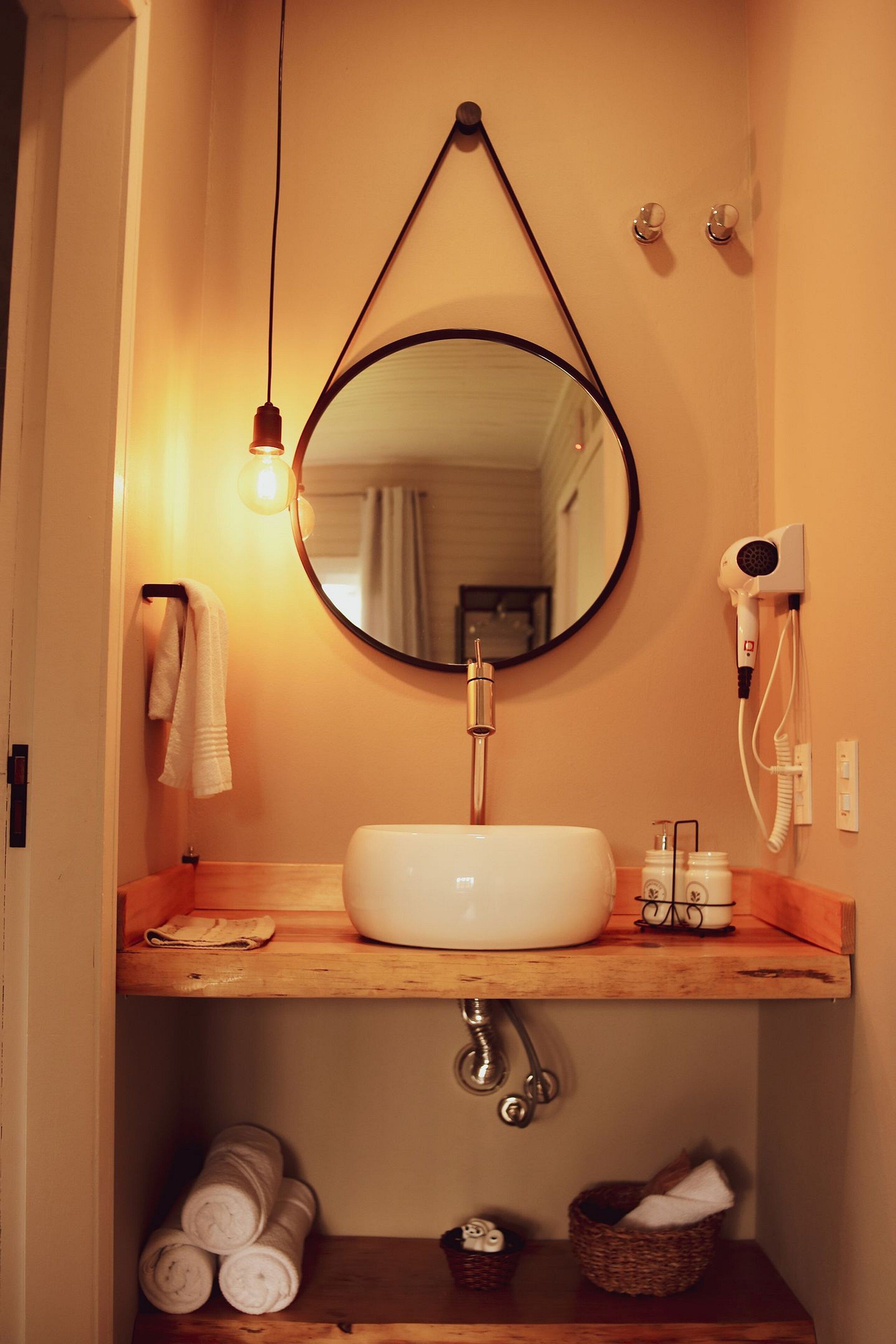 A bathroom with a round mirror, a white sink, a towel rack, a hairdryer, and a wooden shelf.