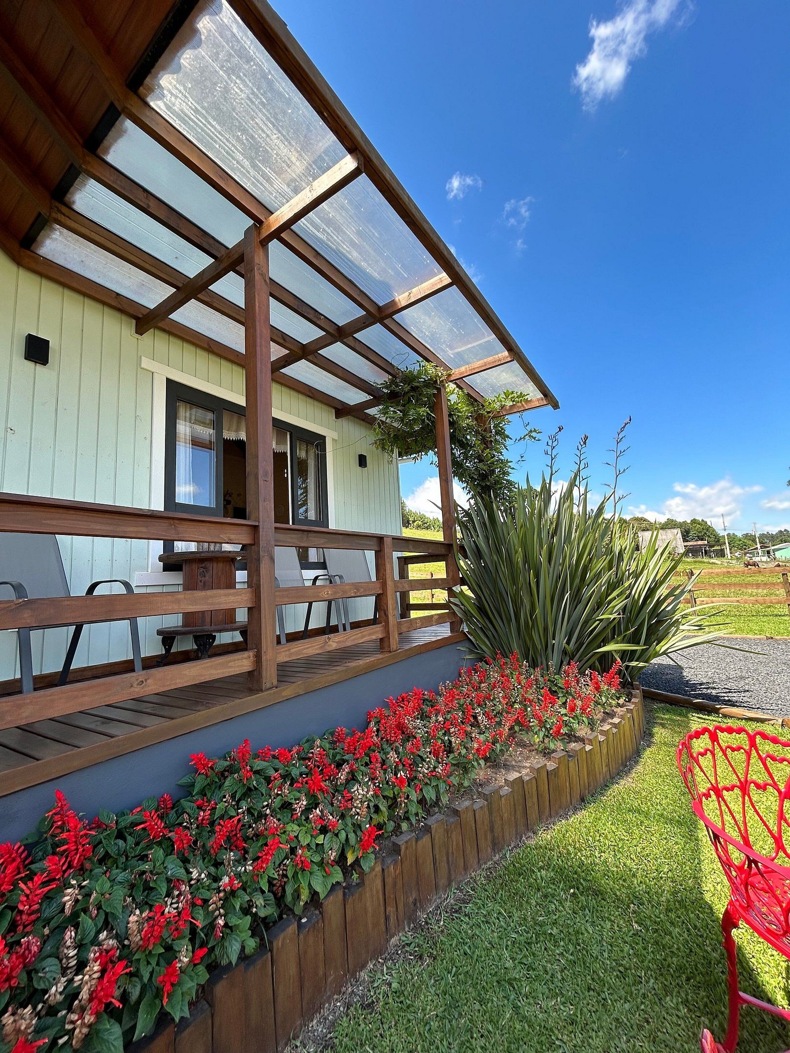 A covered outdoor seating area with wooden benches and a glass roof, surrounded by a flower bed and a lawn.