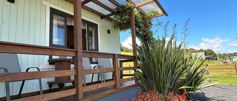 A covered outdoor seating area with wooden benches and a glass roof, surrounded by a flower bed and a lawn.