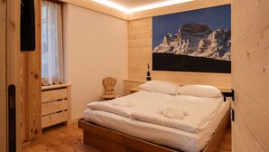 A wooden room with a bed, a window with lace curtains, a painting of mountains, and a wooden nightstand. - Pelmo Mountain Lodge (Borca di Cadore)