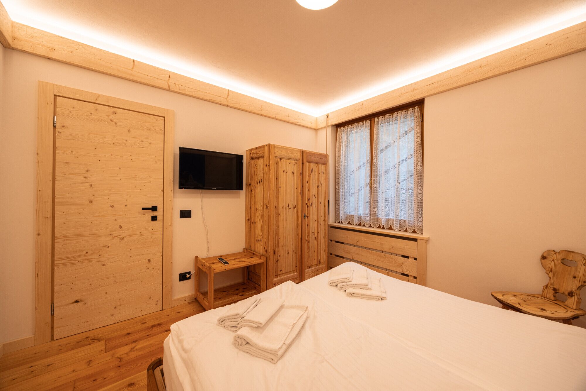 Una habitación de madera con cama, televisor, ventana con cortinas de encaje y armario de madera.