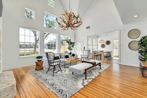 A spacious living room with a high ceiling, large windows, a wooden floor, a sofa, a coffee table, and a chandelier.