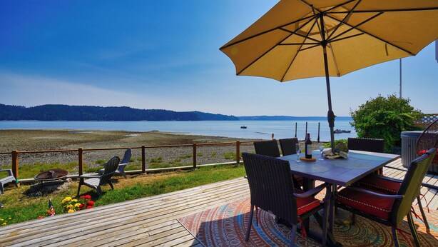 Une terrasse en bois avec des meubles d’extérieur, une table avec une bouteille de vin et une vue sur un lac paisible où flottent des bateaux.