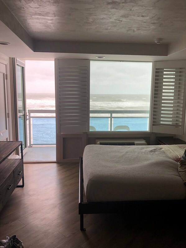 A bedroom with a large bed, a wooden dresser, and a view of the ocean through open shutters.