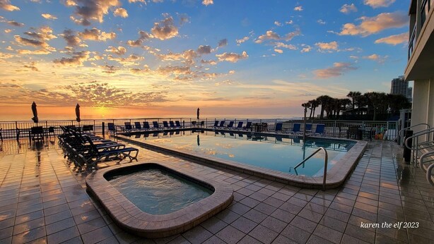 A rooftop pool with lounge chairs and a hot tub, overlooking a sunset and ocean view.