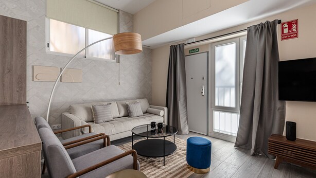Una camera d'albergo moderna con un divano, poltrone, un tavolino, una lampada, una TV e una porta scorrevole che dà su un balcone.