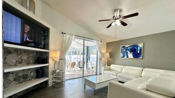 A modern living room with a white sofa, a coffee table, and a TV on a shelf. There is a ceiling fan, a painting on the wall, and a view of the outdoors through large windows. - Comfy Home Near Theme Parks (Kissimmee)