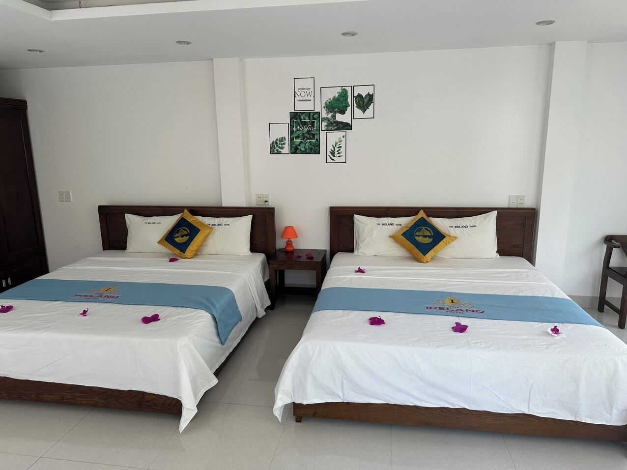 A hotel room with two beds, each with a pillow featuring a logo, a nightstand with a lamp, and a wall decorated with framed pictures.