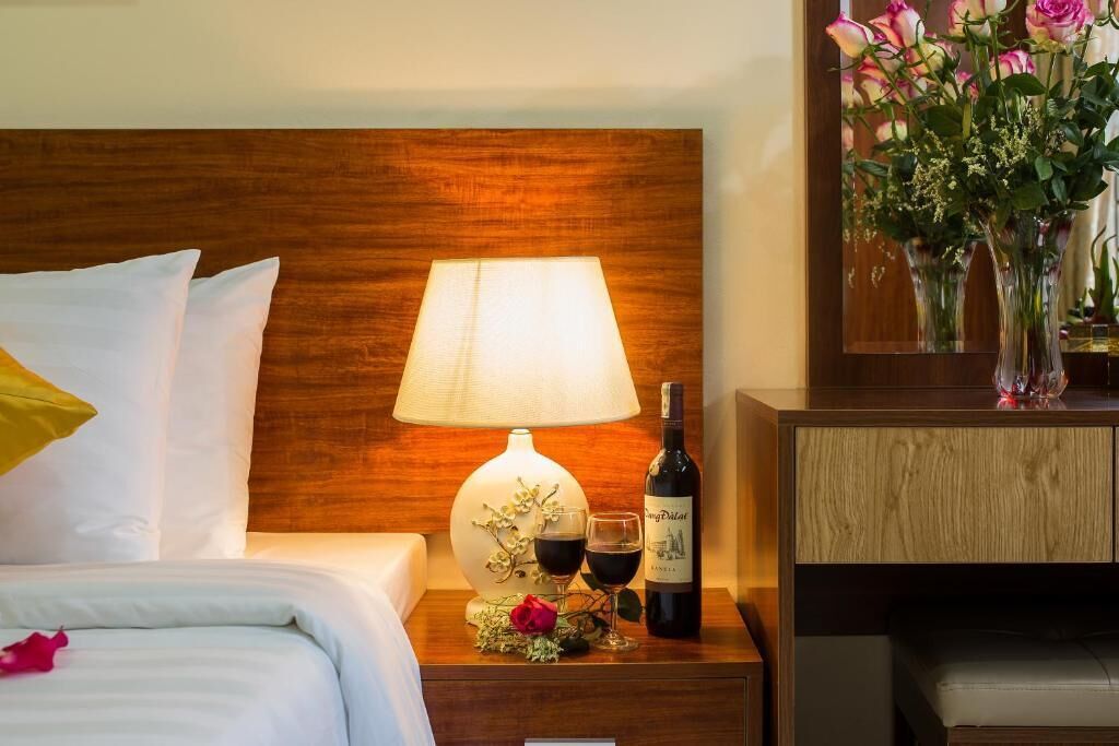 A neatly made bed with white linens, a wooden headboard, a bedside table with a lamp, a bottle of wine, and a vase with flowers.