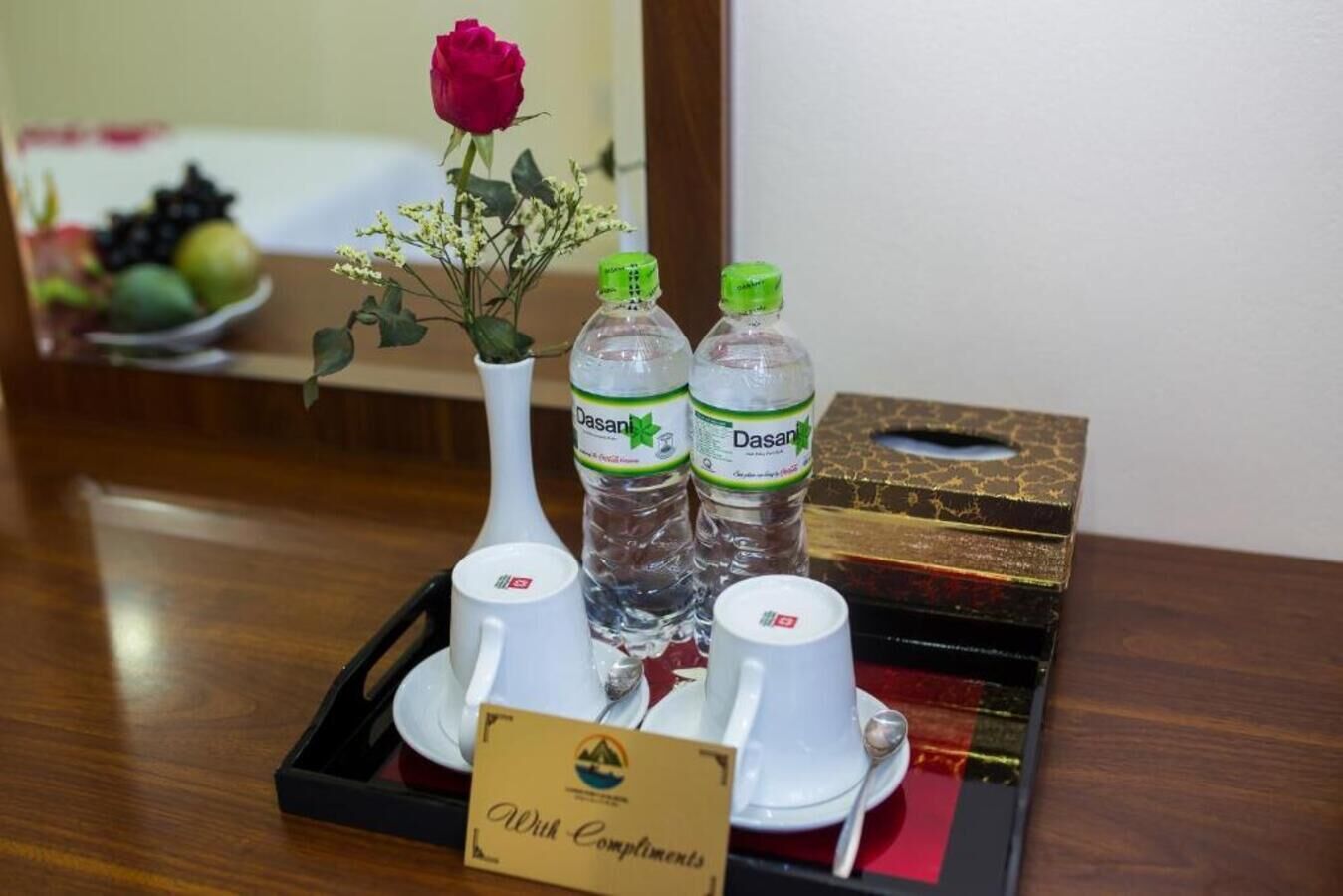 Ein Tablett mit zwei Dasani-Wasserflaschen, einer weißen Vase mit einer roten Rose und zwei weißen Tassen mit Untertassen auf einem Holztisch.
