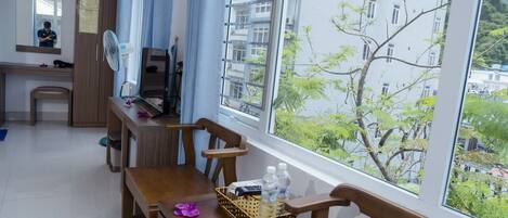 Una habitación con una mesa de comedor de madera, sillas, un escritorio pequeño, una silla, una ventana con vistas a un edificio y árboles, y un ventilador.