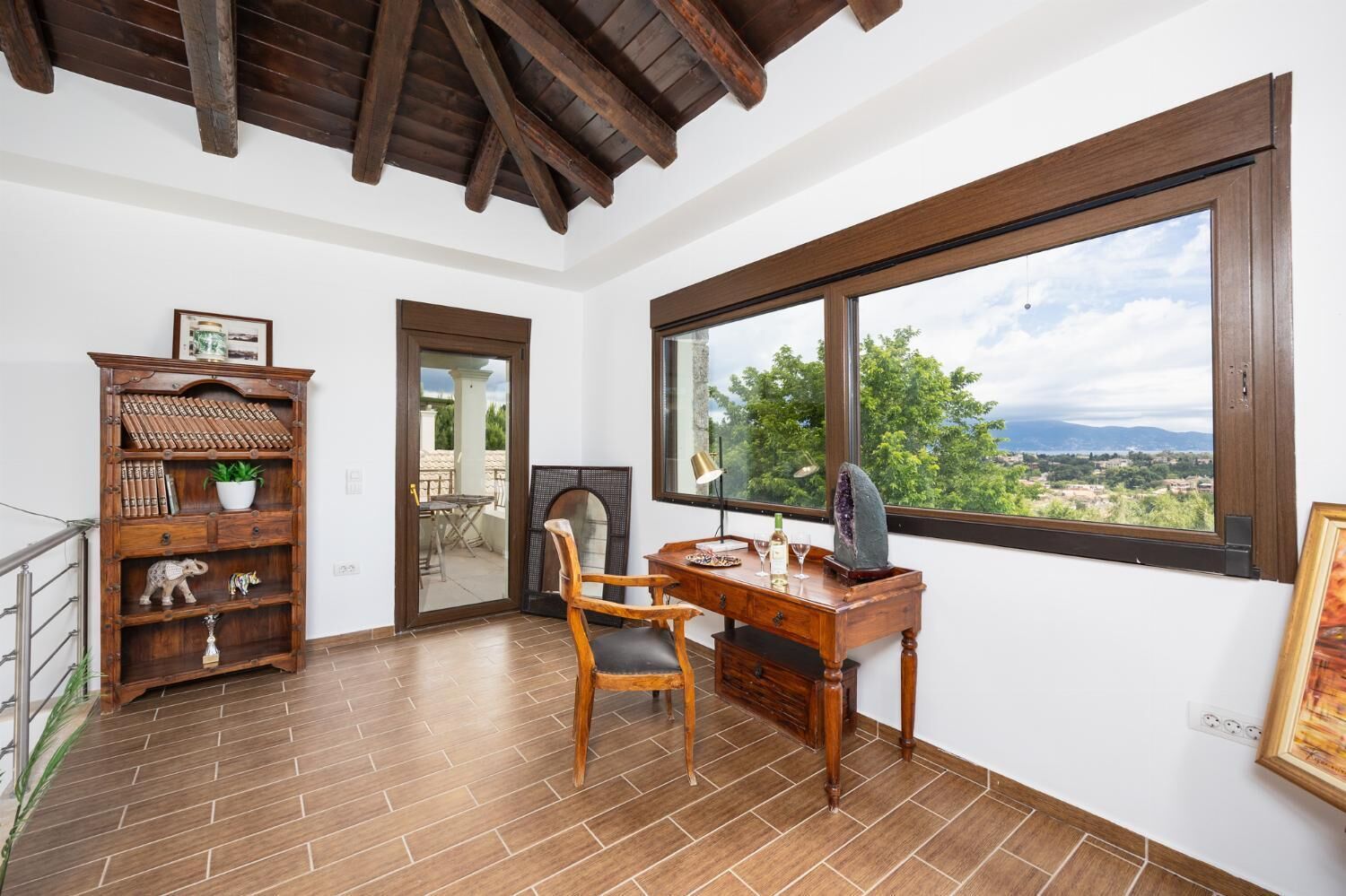 Ein Zimmer mit einem Holztisch, einem Stuhl, einem Bücherregal und einem großen Fenster mit Blick auf Bäume und Berge.