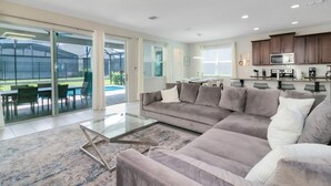 A modern living room with a sectional sofa, a glass coffee table, and a view of a backyard through large sliding glass doors. - Incredible Resort 4 bed Pool Spa Home (Kissimmee)