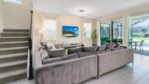 A spacious living room with a large grey sectional sofa, a flat-screen TV mounted on the wall, and a staircase leading to another level. - Incredible Resort 4 bed Pool Spa Home (Kissimmee)