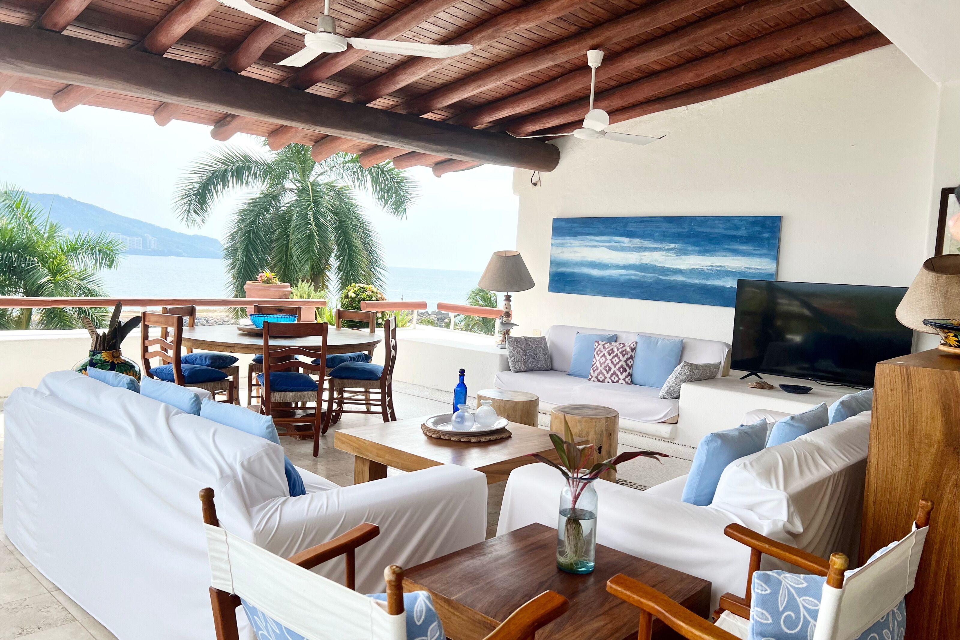A sunlit outdoor seating area with white cushioned sofas and wooden chairs, a wooden table, and a TV on a stand.