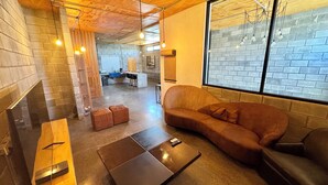 A modern lounge area with a brown sofa, a wooden bench, and a coffee table. There is a brick wall and a large window with a view of the outside.