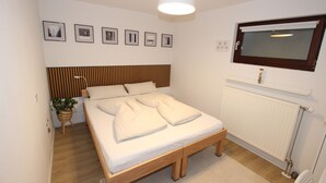 Een slaapkamer met een houten bed, wit beddengoed, een bedlampje, een radiator en ingelijste foto’s aan de muur.