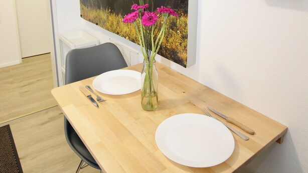 Een eethoek met een houten tafel, twee witte borden, een vaas met roze bloemen en een groot landschap op de muur.