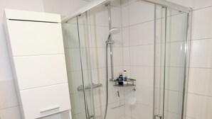 Een douchecabine met glazen wanden, een witte betegelde muur, een wit kastje met lades en een houten vloer.