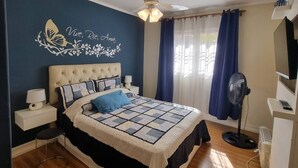 Ein Schlafzimmer mit einem Bett, einem Deckenventilator, einem Nachttisch, Wanddeko und einem Fenster mit Vorhängen.