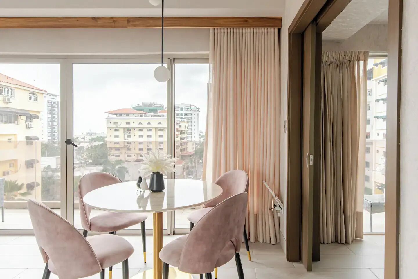 Un espace repas moderne avec une table ronde et quatre chaises, de grandes fenêtres avec des rideaux et une vue sur les bâtiments environnants.