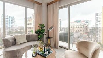 A modern living room with a sofa, a coffee table, and a chair, large windows with curtains, and a view of the cityscape.