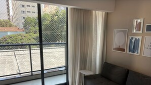 A modern living room with a grey sofa, a small white side table, and a book on a brown ottoman. A large window with sheer curtains offers a view of a cityscape and a fenced outdoor area. - Studio 203 São Paulo prox Allianz Unimed (São Paulo)