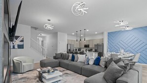 Un salon moderne avec un canapé d’angle gris, un fauteuil blanc et une table basse. On aperçoit une cuisine à l’arrière-plan, avec un mur d’accent bleu.
