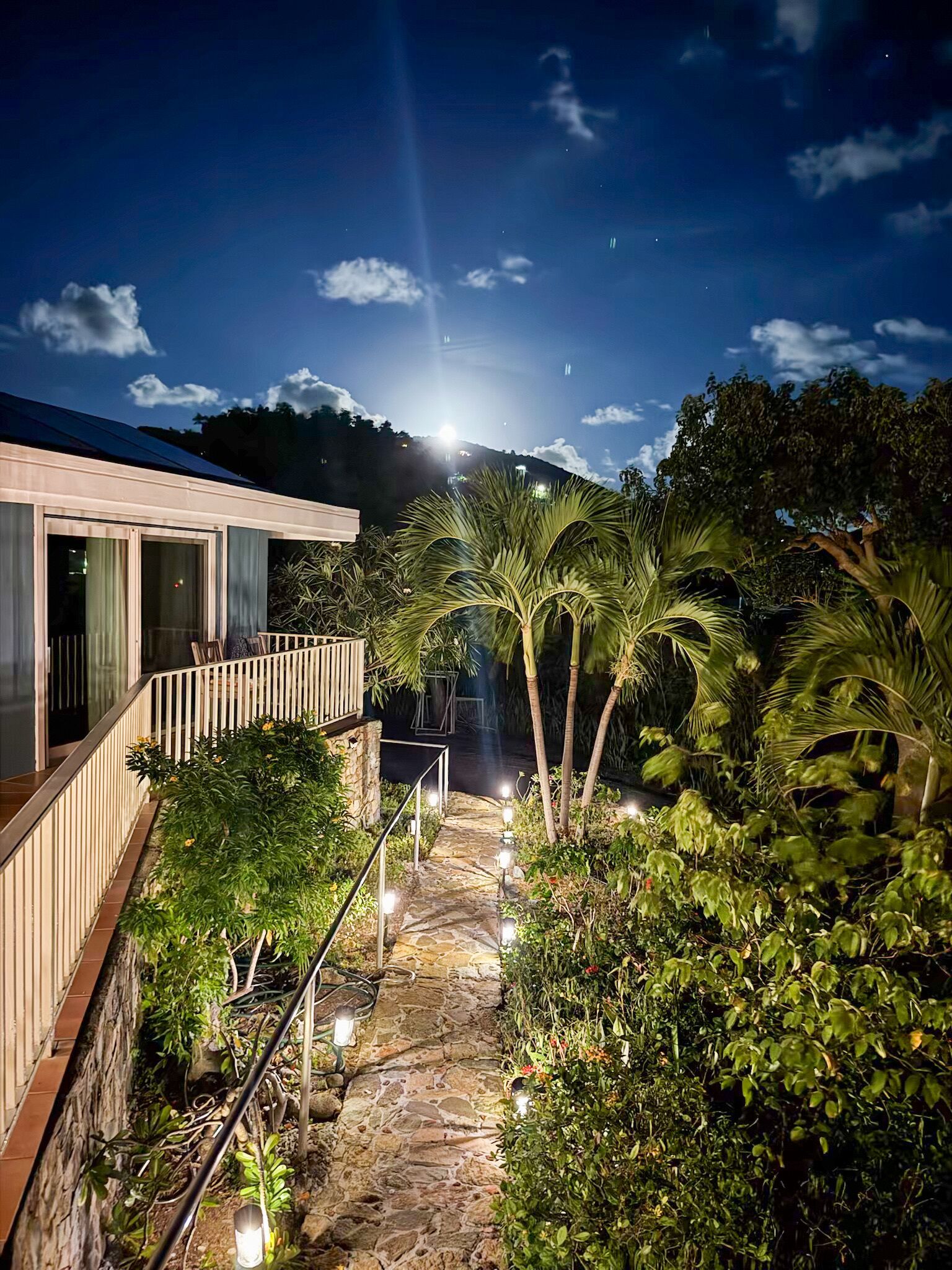 A nighttime view of a house with a balcony overlooking a garden with palm trees and a stone pathway.