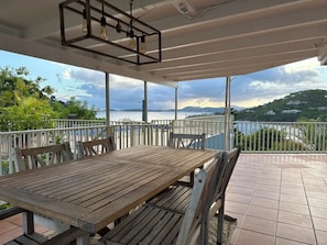 Une table et des chaises en bois, placées sous une terrasse couverte, avec vue sur un plan d’eau et des collines.