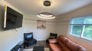 Ein Wohnzimmer mit einem braunen Ledersofa, einer schwarzen Bank und einem schwarzen Hocker. An der Wand ist ein Flachbildfernseher befestigt, eine Deckenleuchte ist installiert, und ein Fenster mit Jalousien vorhanden.
