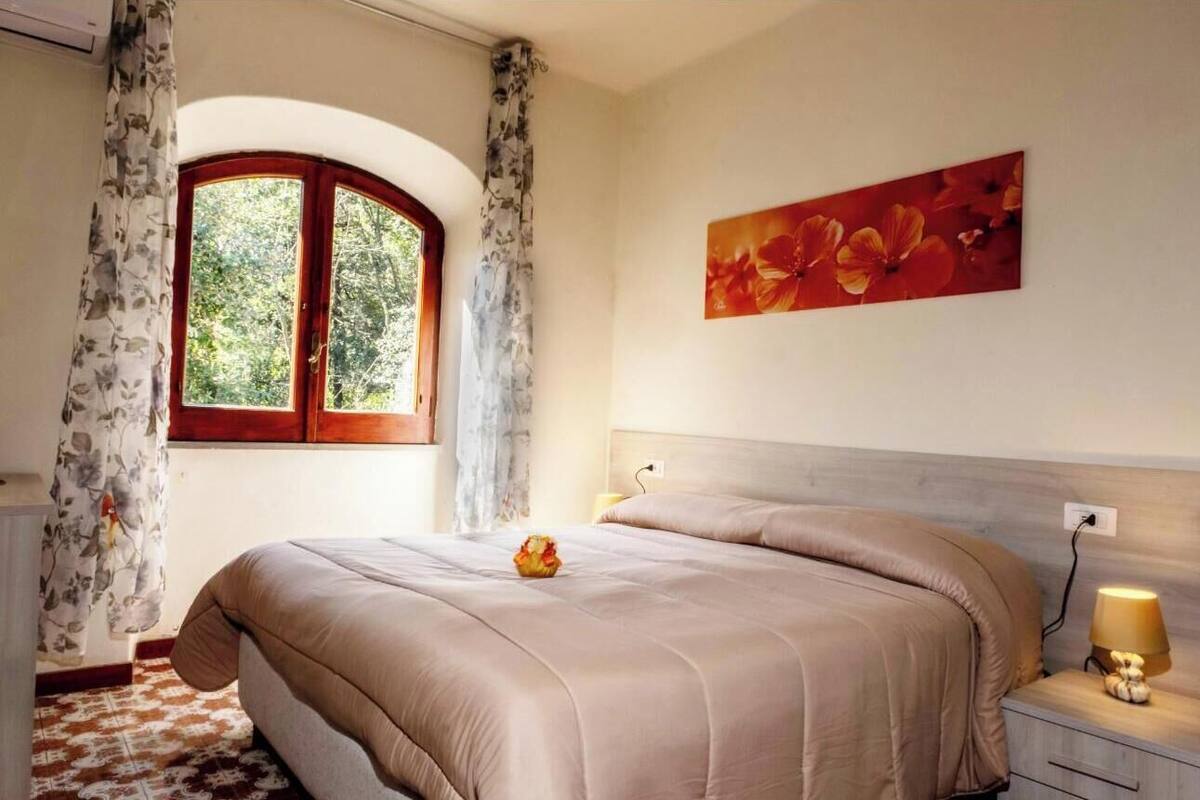Un dormitorio con una cama grande, una ventana con cortinas, una mesita de noche con lámpara y un cuadro floral en la pared.