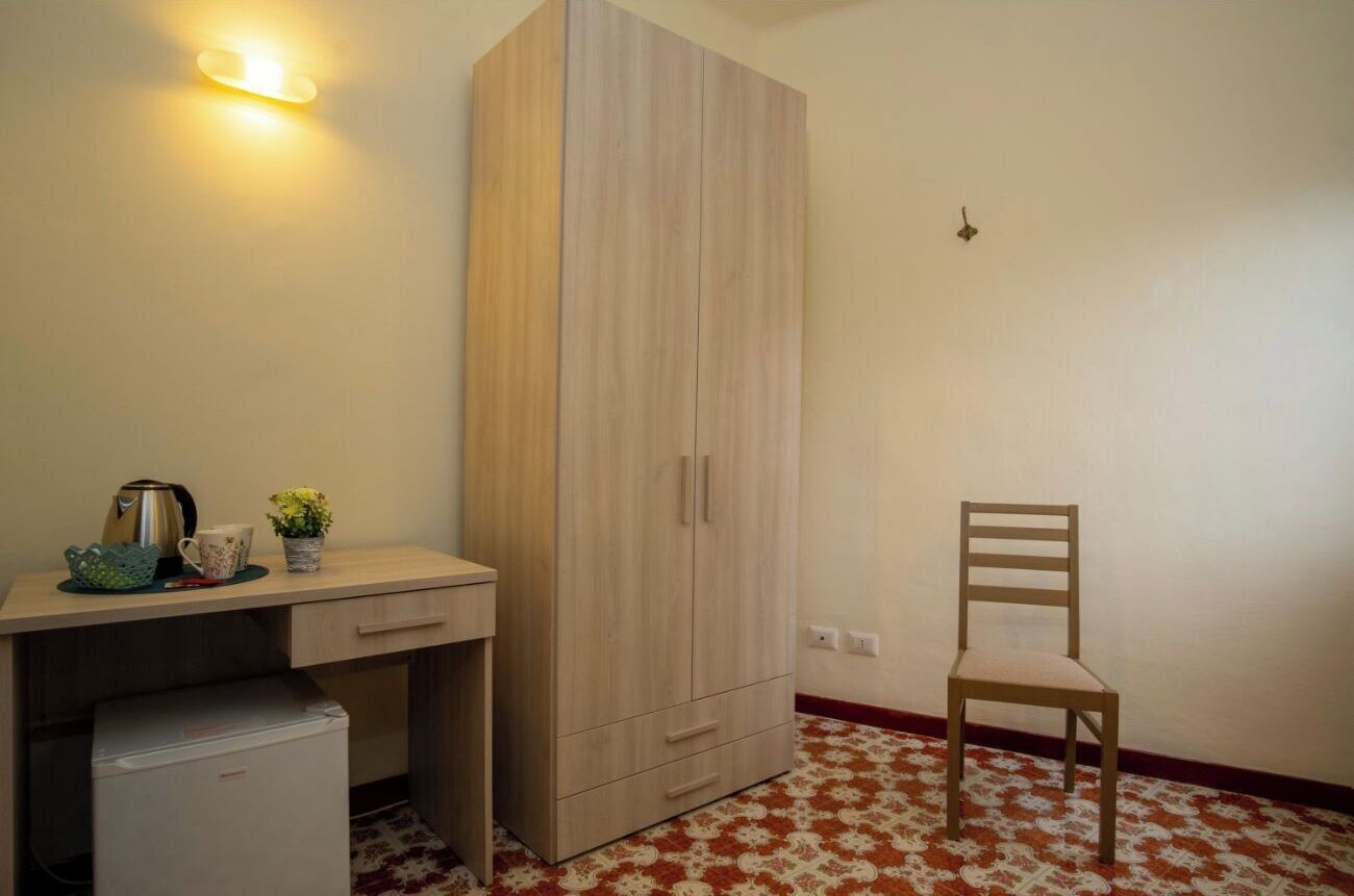 A room with a wooden wardrobe, a small desk with a kettle and a cup, a chair, and a patterned floor.