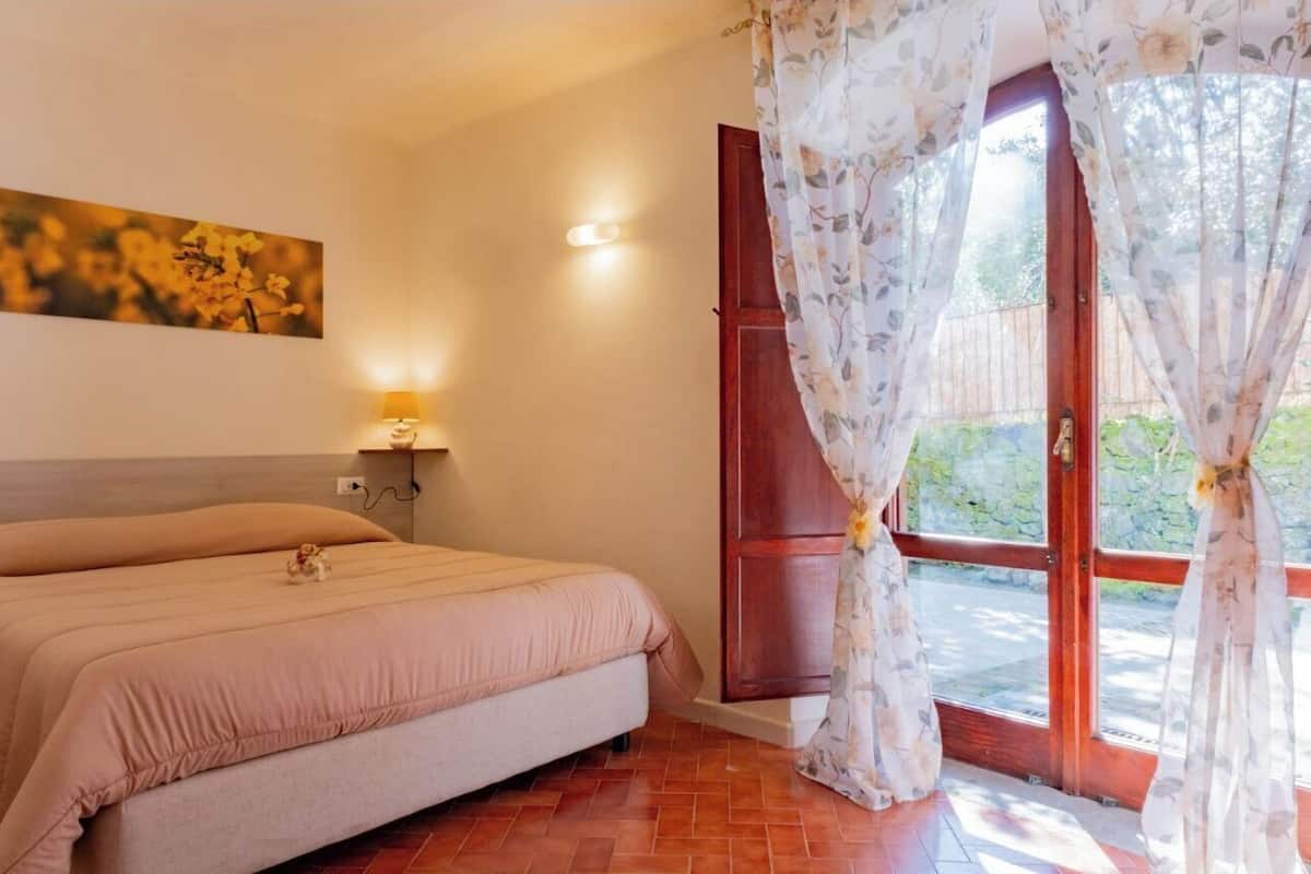 Un dormitorio con cama, mesita de noche, lámpara, un cuadro en la pared y ventana con cortinas.