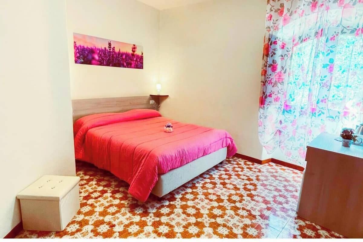 Un dormitorio con cama, mesita de noche, lámpara de pared, una pequeña mesa auxiliar y suelo estampado.