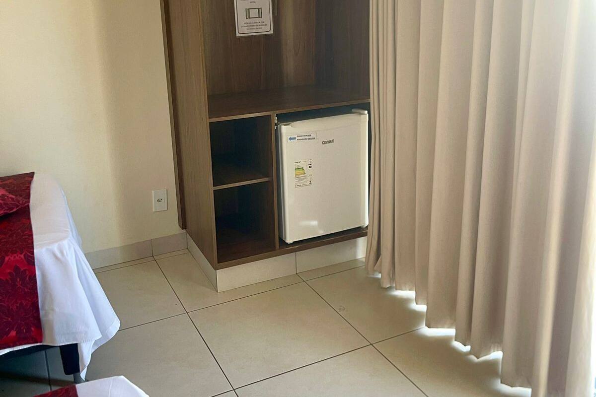 A hotel room with a wardrobe, a small refrigerator, a bed with a red and white bedspread, and a curtain.