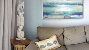 A living room with a beige sofa, a decorative wooden sculpture, a cushion with a fish pattern, and a painting of a seascape. - Isle B Back Seawall Gulf & Pool View! Come Chillax (Galveston)