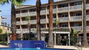A swimming pool with blue tiles, surrounded by palm trees and a multi-story hotel building with balconies. - Beach Escape Seawall View 2 Pool (Galveston)