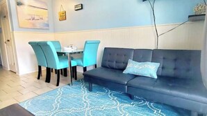 A room with a dark grey sofa, two turquoise chairs, a small round table, a blue patterned rug, and a wall-mounted TV. - Beach Escape Seawall View 2 Pool (Galveston)