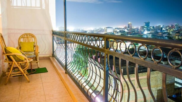 Un balcón con barandilla metálica curva, vista a un paisaje urbano al anochecer y dos sillones de mimbre con cojines.