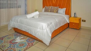Una cama bien tendida con una colcha blanca y almohadas, una mesita de noche de madera con una lámpara, y un tapete floral colorido.