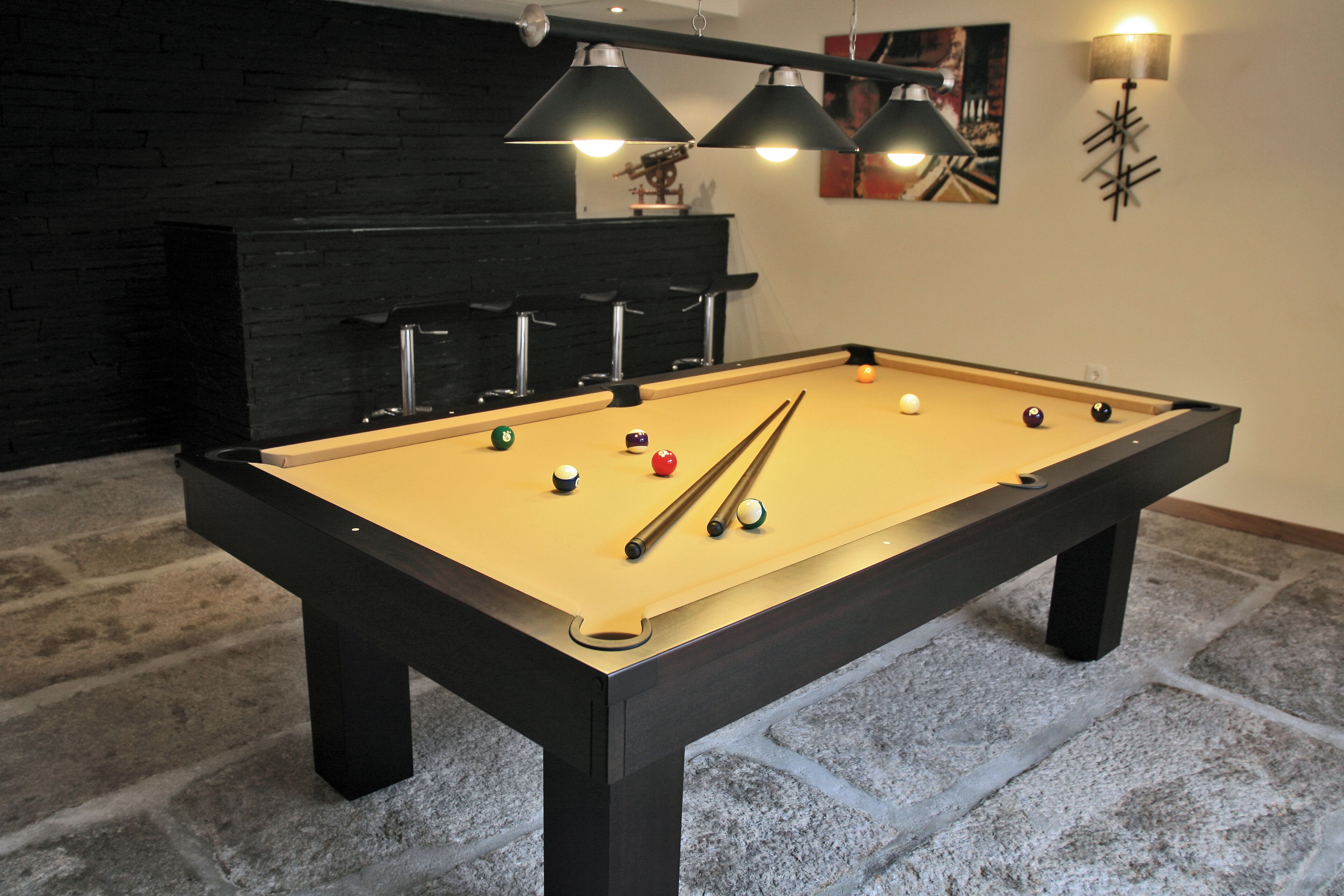 Una mesa de billar con bolas y tacos, una pared negra con chimenea y una lámpara moderna.