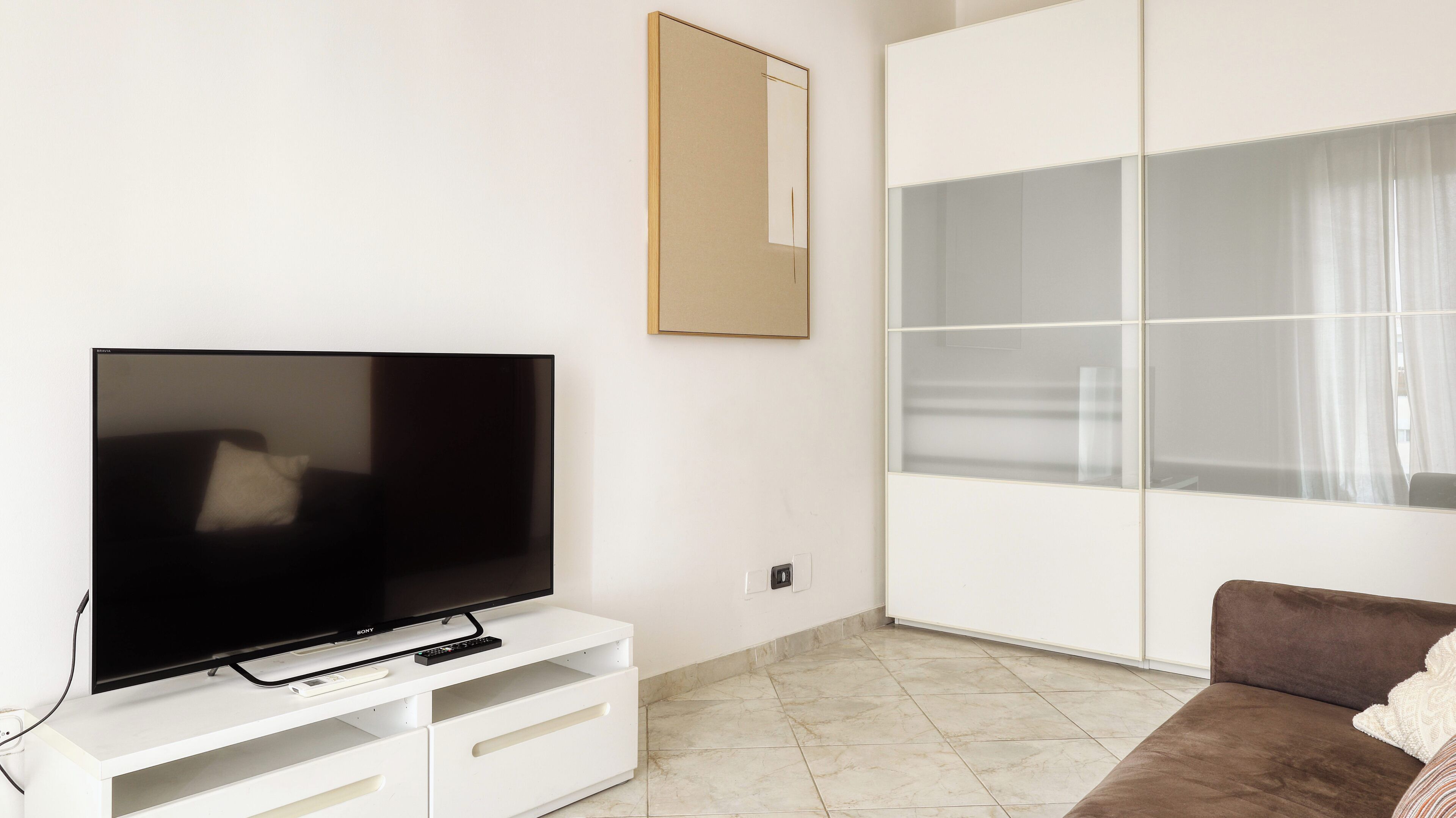 A modern living room with a flat-screen TV on a white entertainment unit, a brown sofa, and a large sliding glass door leading to a balcony.