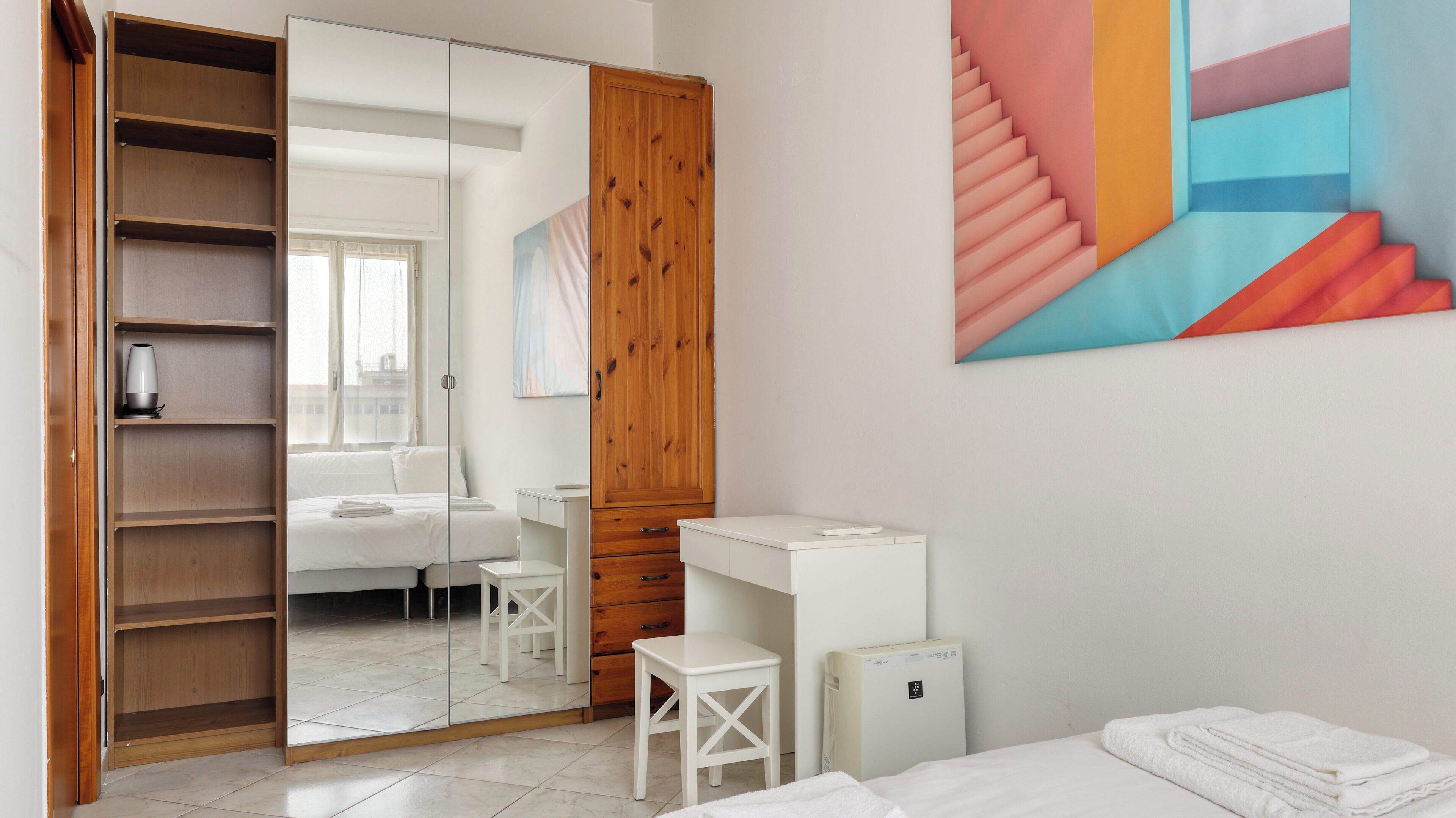 A bedroom with a wooden wardrobe, a white desk, a bed, and a large mirror.